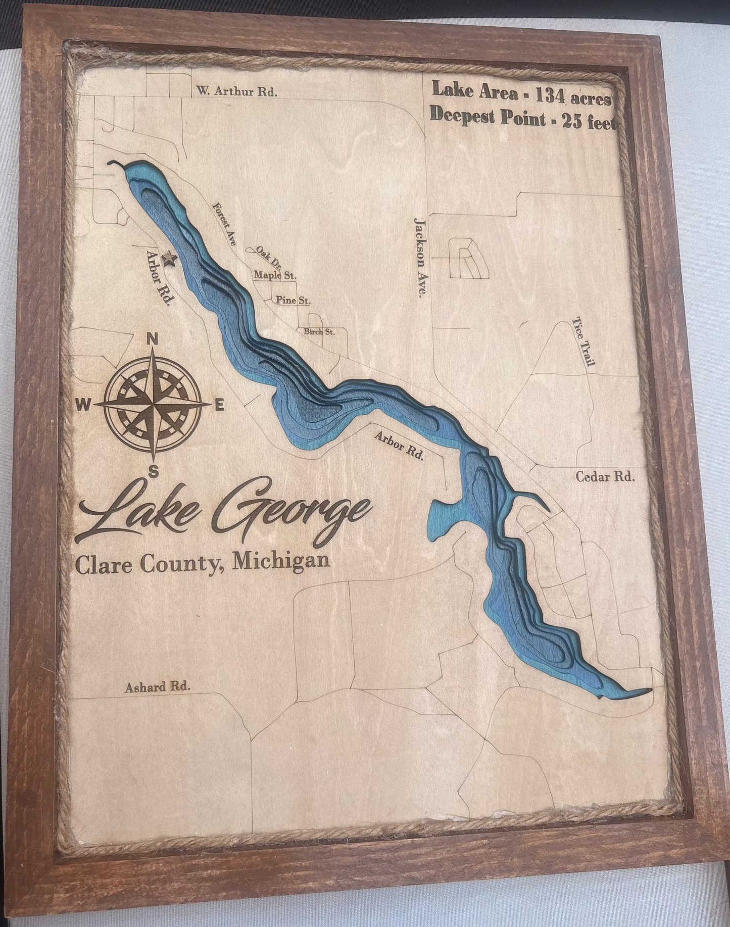 A framed wooden art piece depicting a vintage-style map of Lake George in Clare County, Michigan, with blue water channels and black text details, set against a neutral background.